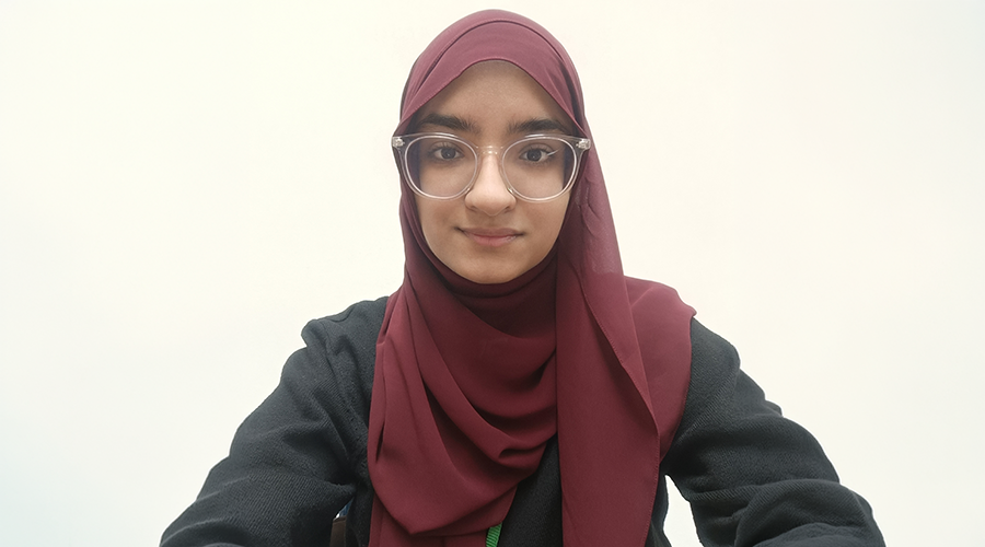 Maryam Ilyas in glasses and a maroon hijab is facing the camera and smiling slightly. The background is plain and light-colored.