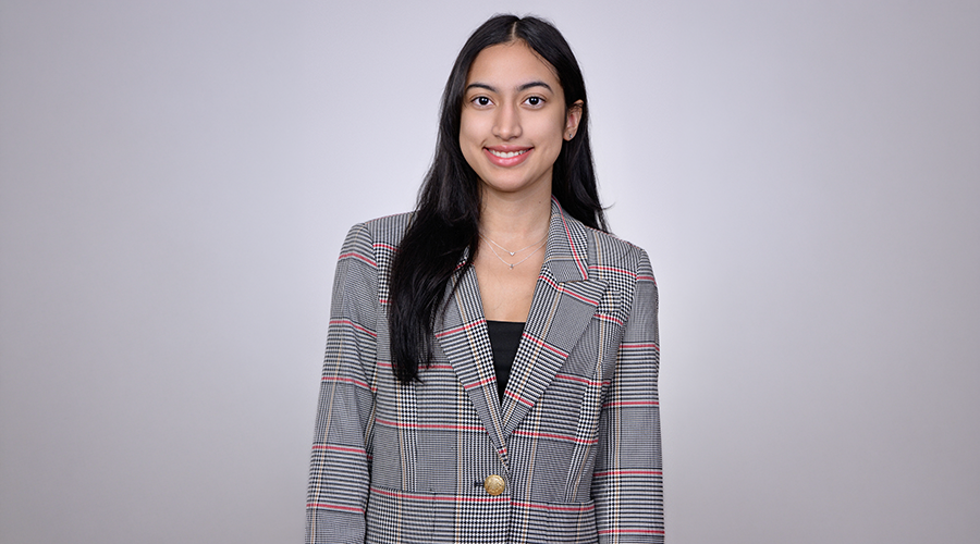 Pamela Rivera in long dark hair smiles, wearing a plaid blazer with a gold button over a black top, standing against a plain light gray background.