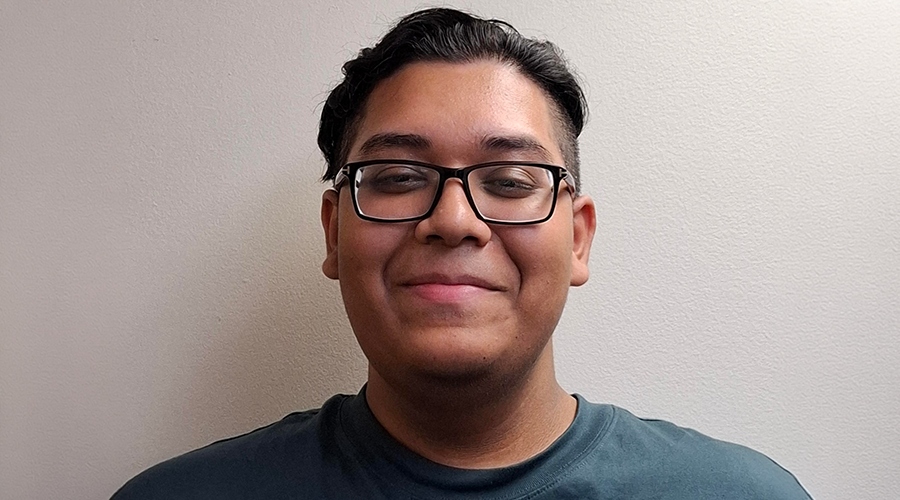 Shazadul Islam with short dark hair and glasses smiles slightly while standing in front of a plain, light-colored wall, wearing a teal t-shirt.