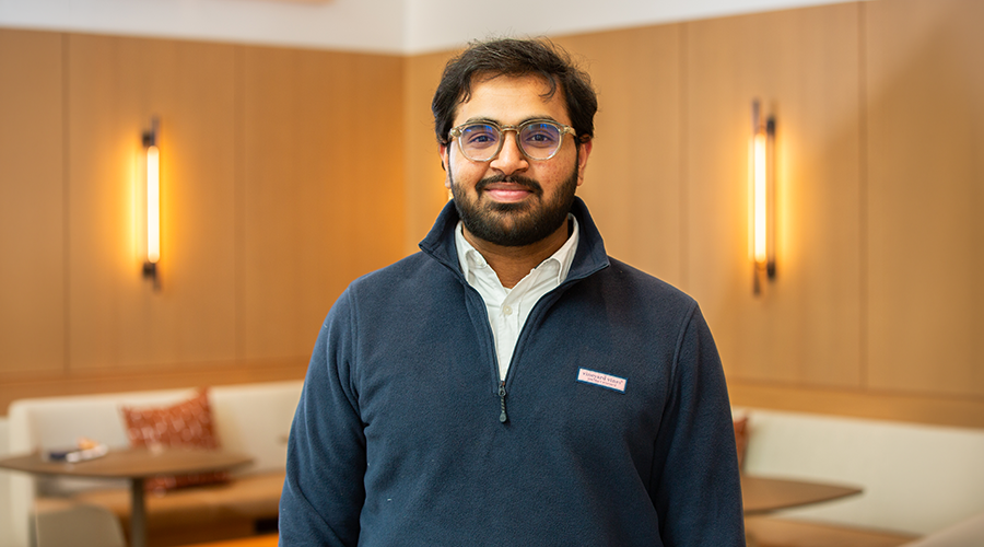 Subhan Tariq with glasses and a beard, wearing a navy pullover and white collared shirt, stands indoors in a warmly lit room with light wooden walls and cushioned seating in the background.