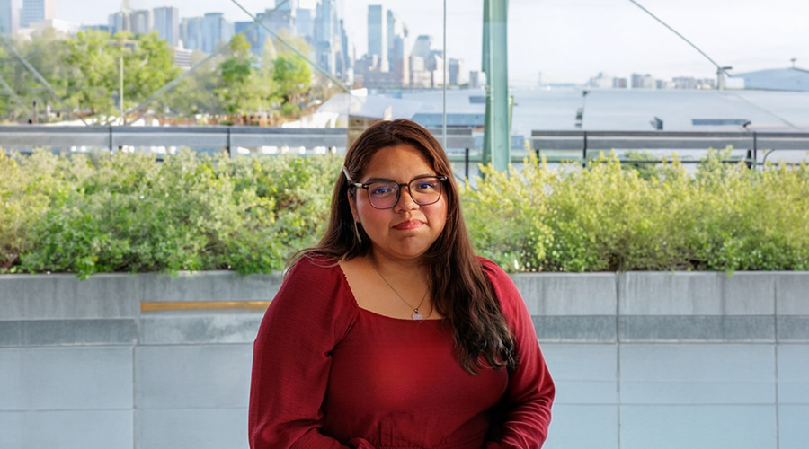 Yadhira Vivaldo Garcia with long brown hair and glasses, wearing a red blouse, sits outdoors in front of greenery and glass windows, smiling slightly at the camera.