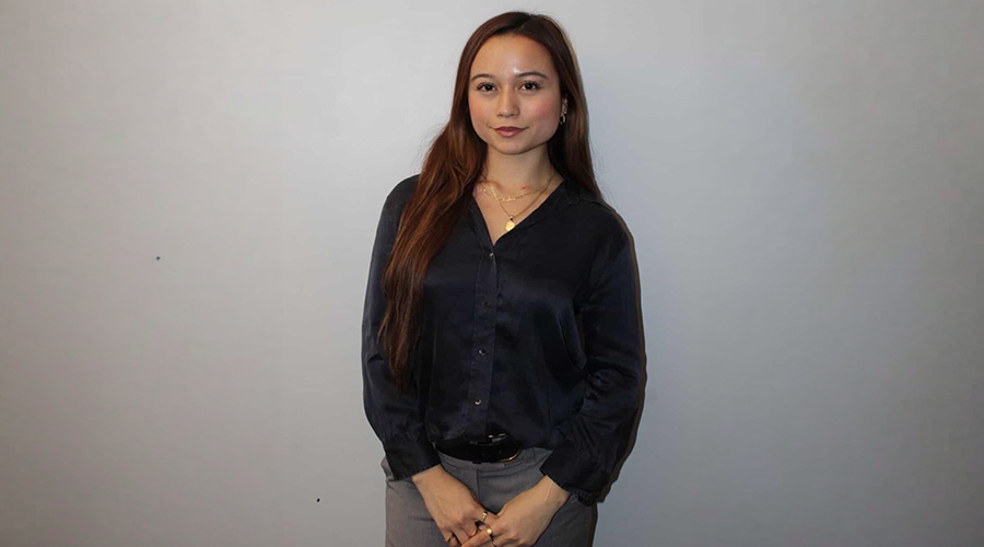 Andrea Quinonez with long brown hair, wearing a dark blouse and gray pants, stands against a plain light gray background with her hands clasped in front of her, looking at the camera.