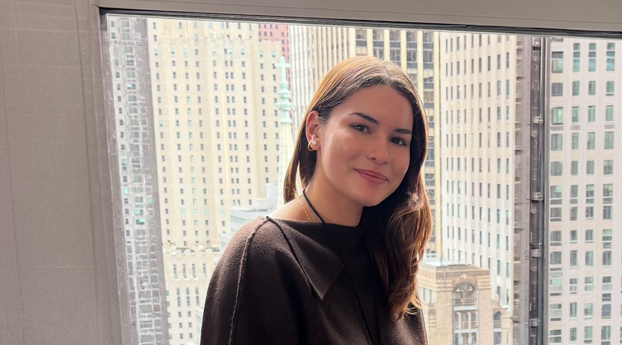 Mariana Gonzalez with long brown hair, wearing a dark top, smiles while sitting indoors in front of a large window with a New York City skyline of tall buildings in the background.