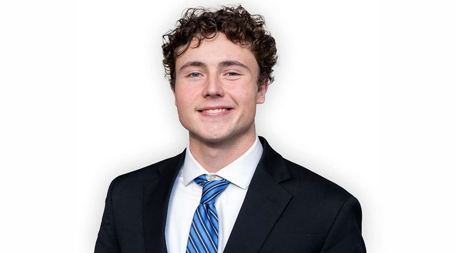 Ron Kullashi with curly brown hair, wearing a black suit, white shirt, and blue striped tie, smiles at the camera against a plain white background.