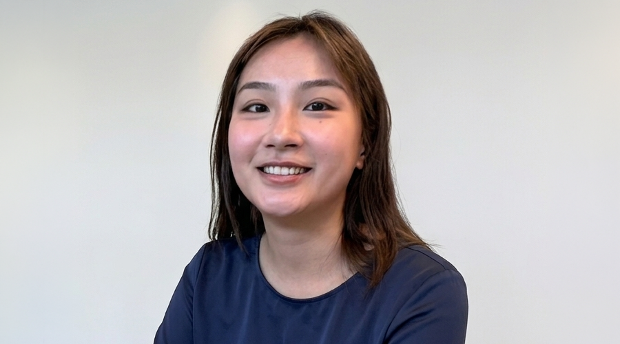 Shasuna Lin with straight brown hair and a navy blue shirt smiles at the camera against a plain, light-colored background.