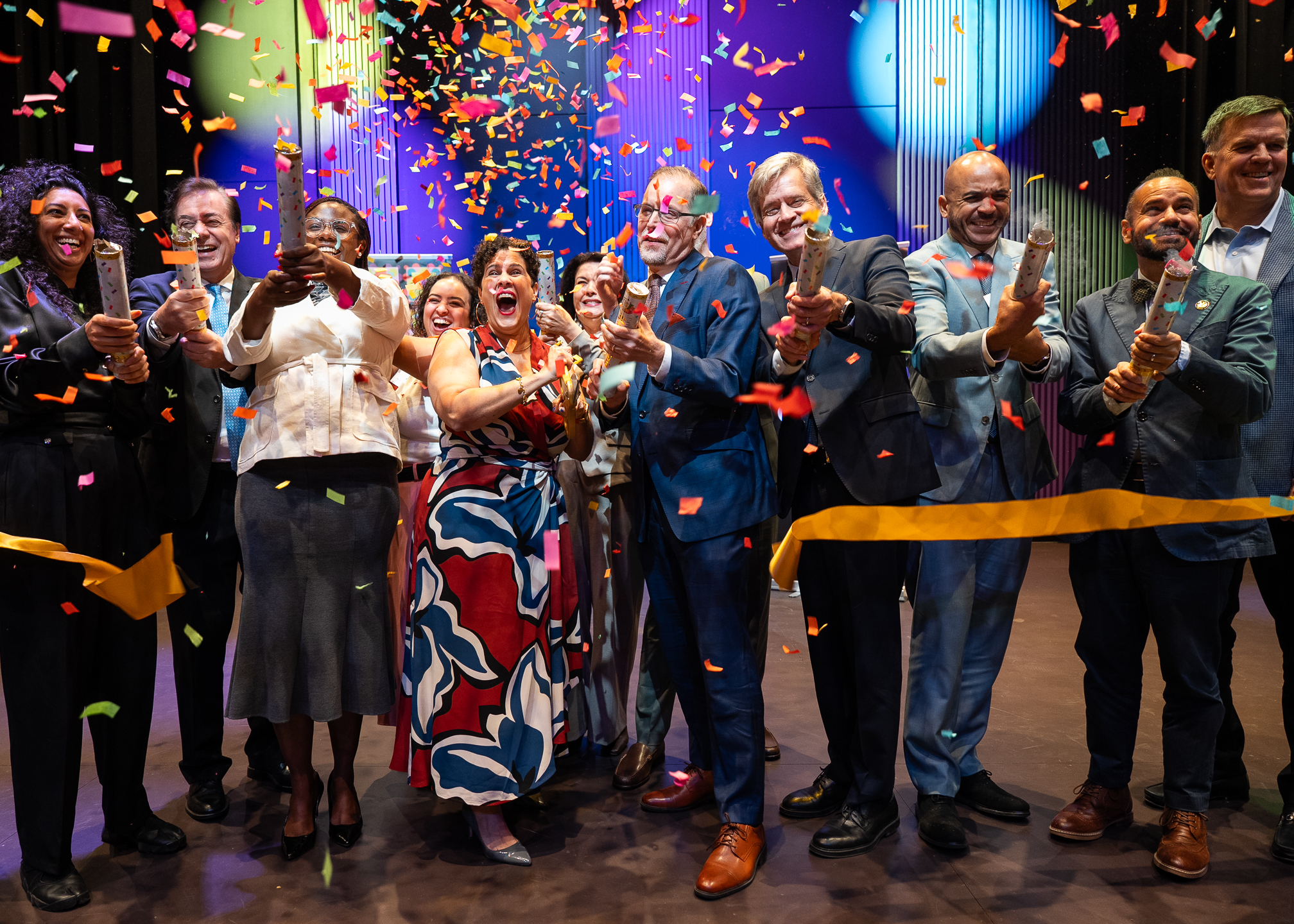A group of people celebrates at a ribbon-cutting ceremony, releasing colorful confetti. They are smiling, clapping, and dressed in formal attire on a decorated stage.