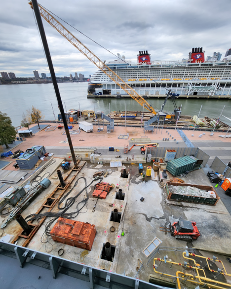 A construction site near a river with equipment, tools, a crane, and a large cruise ship docked in the background on a cloudy day.