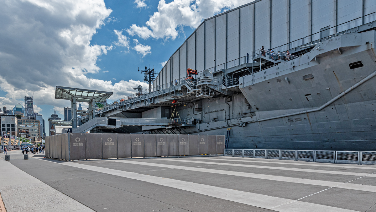 A large gray aircraft carrier is docked near a city with people walking by, a tall fence, and buildings visible in the background under a blue sky with clouds.