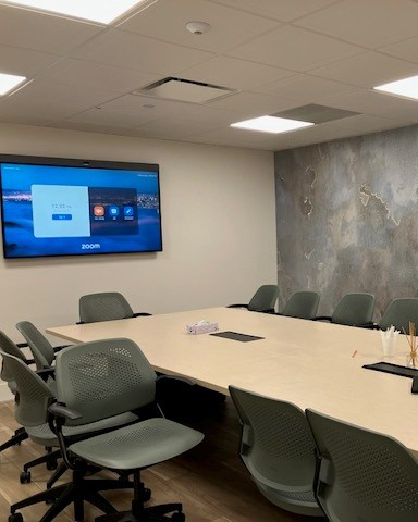 A modern conference room with a long table, several green chairs, a large wall monitor displaying a Zoom screen, and a textured accent wall in the background.