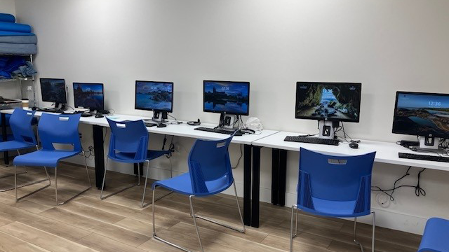 A row of five desktop computers with monitors sits on white tables against a wall, with blue chairs in front of each station. The room has wood flooring and a few blue mats stacked in the corner.