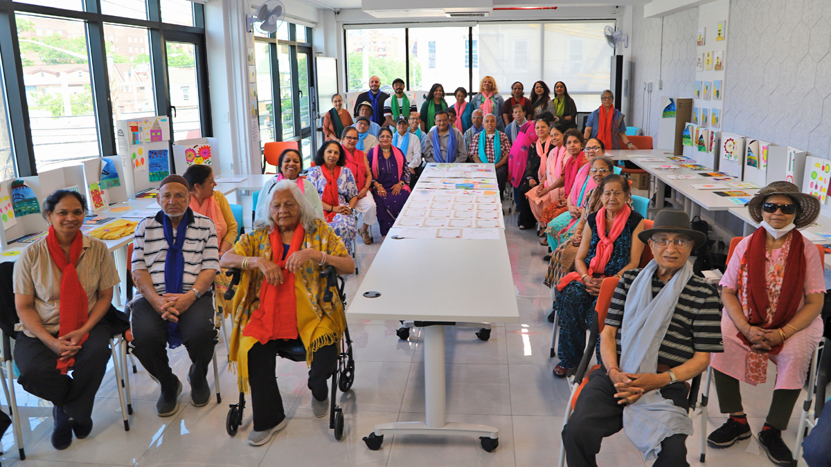 A group of seniors and adults pose in a bright classroom with artwork displayed on the walls and tables. Some are seated, others standing, all smiling and wearing colorful scarves.