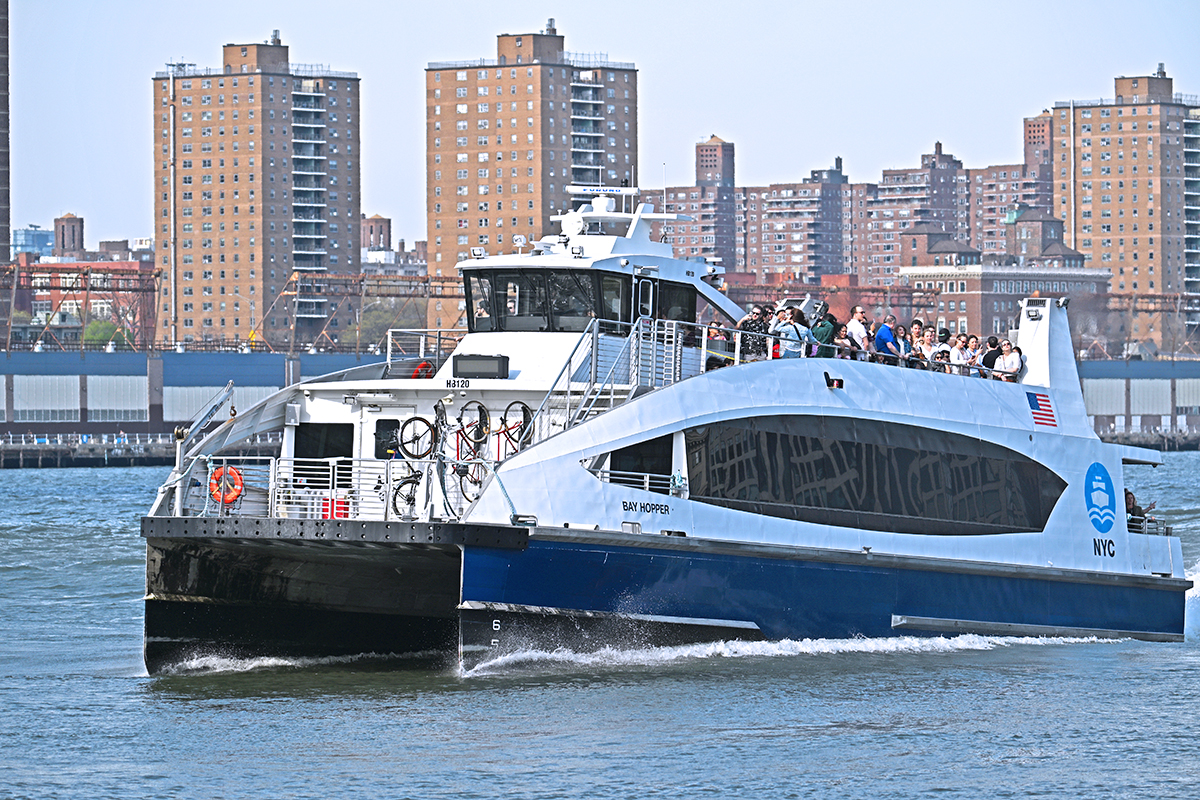 A blue and white NYC Ferry boat, the Bay Hopper, travels across the water with passengers on the upper deck and bicycles racked on the front.