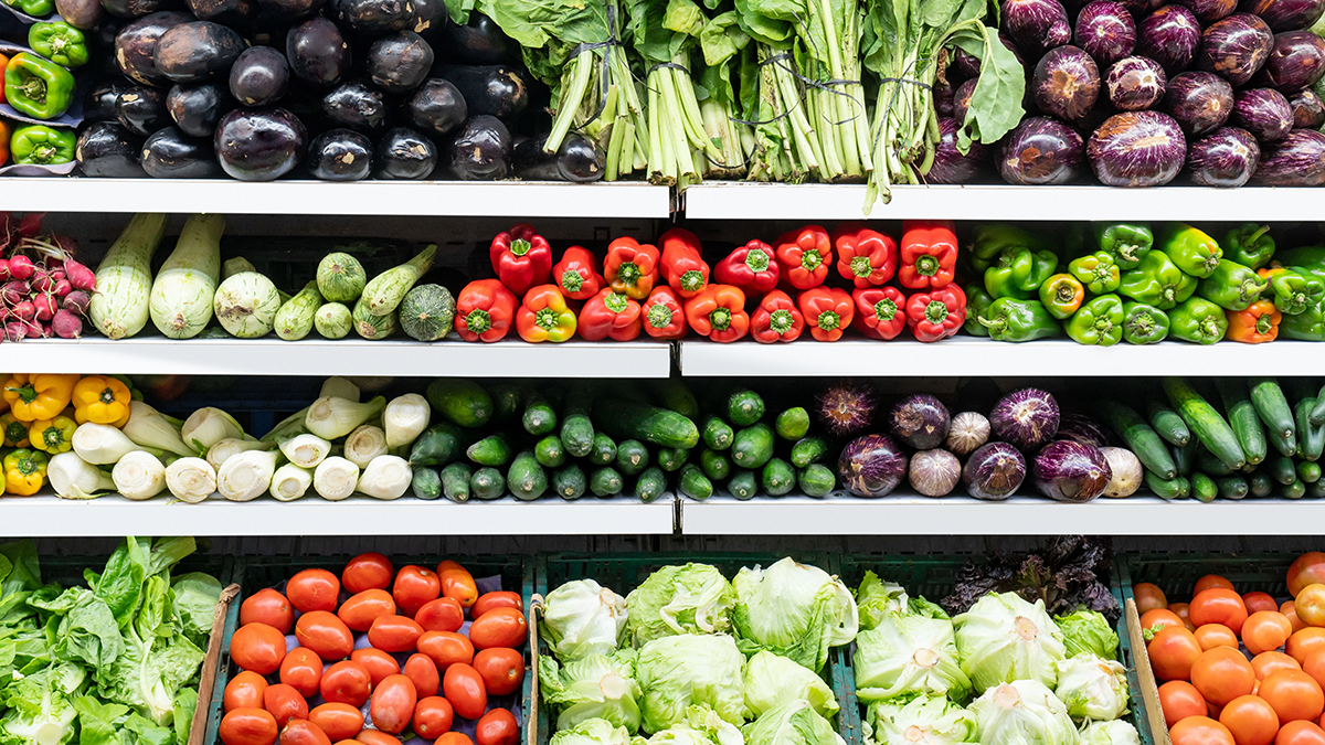 Shelves of fresh vegetables in a grocery store, including eggplants, leafy greens, peppers, cucumbers, tomatoes, and cabbages, all neatly arranged in rows.