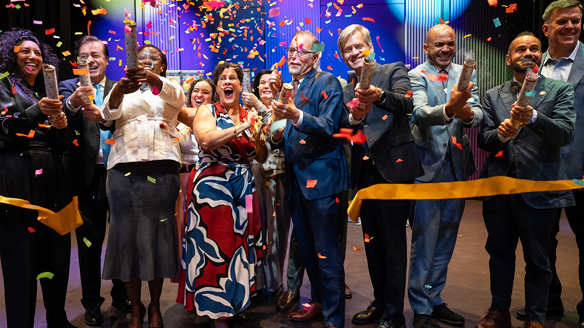 A group of people celebrates at a ribbon-cutting ceremony, releasing colorful confetti. They are smiling, clapping, and dressed in formal attire on a decorated stage.