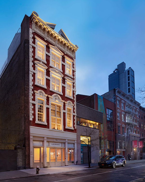 A tall, narrow red brick building with ornate white trim is lit warmly at dusk. The ground floor has large windows and a sign reading La MaMa. Cars are parked along the street with other buildings visible nearby.