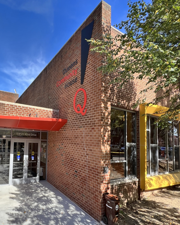 A modern brick building with large windows and a bold red entrance canopy. The sign reads “Queens Community House” with a large red “Q” logo. Trees partially shade the building under a clear blue sky.
