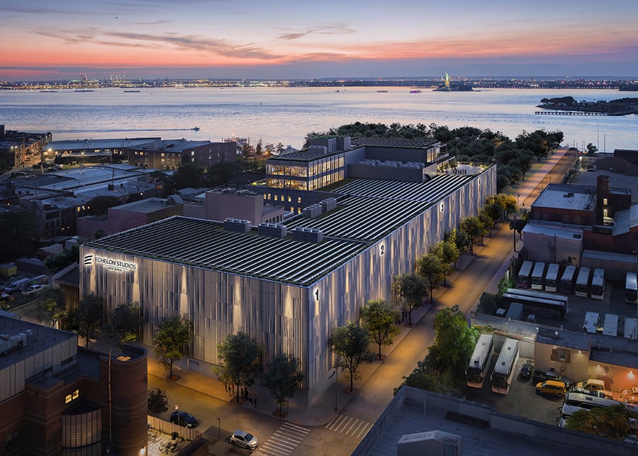 A modern building with a solar-paneled roof is surrounded by trees and city streets at sunset, overlooking a body of water with the Statue of Liberty visible in the distance.