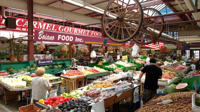 A bustling scene at Arthur Avenue Market with two people shopping for fresh produce. Colorful fruits and vegetables like tomatoes, avocados, and zucchinis fill the stalls. Overhead, a large decorative wagon wheel and market signs are visible.