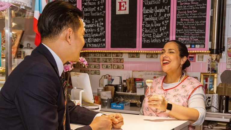 A man in a suit leans on a counter, smiling at a woman in a pink apron holding a dessert. Behind her, a chalkboard menu is visible. The setting is colorful and lively, with various decorations and small framed pictures on the wall. A man in a suit leans on a counter, smiling at a woman in a pink apron holding a dessert. Behind her, a chalkboard menu is visible. The setting is colorful and lively, with various decorations and small framed pictures on the wall.