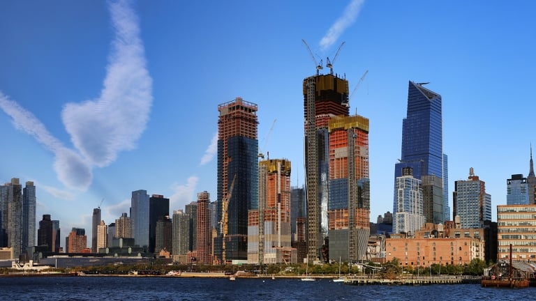 Panoramic view of the New York City skyline featuring modern skyscrapers of Hudson Yards under construction against a clear blue sky. A river is in the foreground, and fluffy white clouds add to the scenic urban landscape.