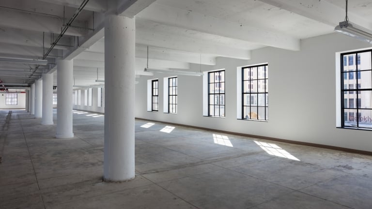Spacious industrial-style interior of Brooklyn Army Terminal with concrete floors and large white pillars. Sunlight streams through multiple large windows along the walls, creating rectangular patterns on the floor. Exposed ceiling pipes add to the minimalist aesthetic. Spacious industrial-style interior of Brooklyn Army Terminal with concrete floors and large white pillars. Sunlight streams through multiple large windows along the walls, creating rectangular patterns on the floor. Exposed ceiling pipes add to the minimalist aesthetic.