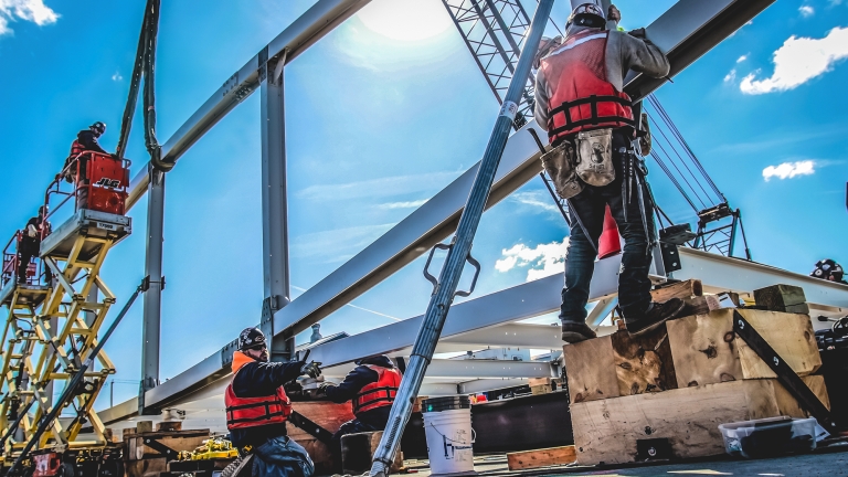 Construction workers in safety gear assemble a metal structure under a clear blue sky. A crane and a scissor lift are in use, with sunlight creating a bright and dynamic atmosphere.