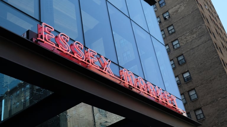 Exterior view of Essex Market with a red neon sign on a modern glass building. A brick building is partially visible in the background, under a cloudy sky.