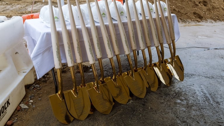 A row of gold-colored ceremonial shovels from the 168th Street ground breaking with light handles is lined up against a table covered with a white cloth. Hard hats are placed on the table, and construction materials are visible in the background.