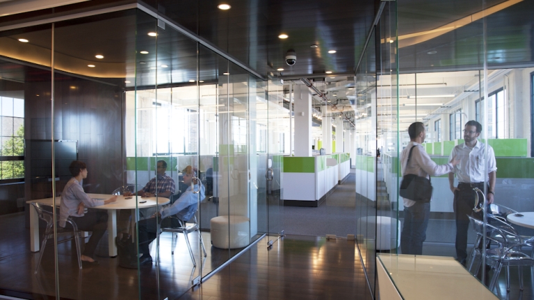 Modern office with glass walls and partitions. Employees are sitting at a round table and others are walking past outside. Green and white color scheme with large windows letting in natural light.