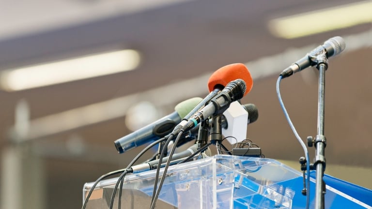 Multiple microphones in various colors are set up on a clear podium, ready for a speech or press conference. The background is blurred, focusing attention on the microphones.