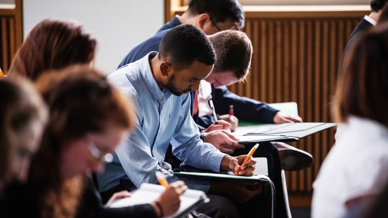 A group of LifeSci NYC applicants sitting at desks in a classroom, focused on writing in notebooks. The students are diverse, dressed in casual attire, and are positioned in rows. The setting is well-lit, with wooden paneling on the walls. Photo by Jordan Rathkopf/NYCEDC.