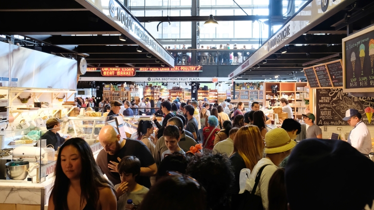 Essex Market, a bustling indoor market with many people browsing various stalls, is well-lit with overhead signage displaying vendor names. Shoppers of diverse backgrounds move through the crowded aisles under a high ceiling with large windows. Essex Market, a bustling indoor market with many people browsing various stalls, is well-lit with overhead signage displaying vendor names. Shoppers of diverse backgrounds move through the crowded aisles under a high ceiling with large windows.