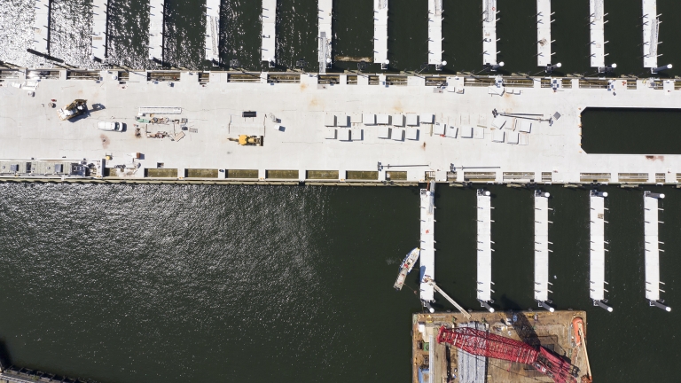 Aerial view of Brooklyn Navy Yard Ferry Pier with numerous empty docks extending into the water. A large pier runs horizontally across the center, with scattered vehicles and equipment on it. The water appears dark, contrasting with the white docks.