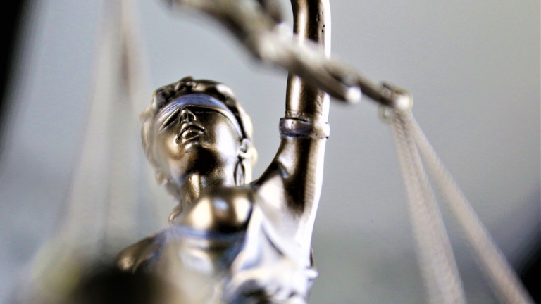 Close-up of a metallic statue of Lady Justice, featuring a blindfolded woman holding scales. The focus is on her face and raised arm, with the background softly blurred.