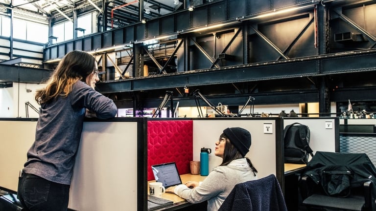 Two individuals in a modern office with high ceilings and industrial design. One person is seated at a desk with a laptop, and the other leans against the partition. The space has exposed beams and large windows.