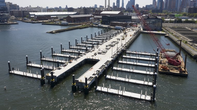 Aerial view of the Brooklyn Navy Yard Ferry pier construction with an L-shaped dock structure extending into the water. Several empty dock slips are visible. A red crane and construction materials are on-site. Cityscape and industrial buildings are in the background.