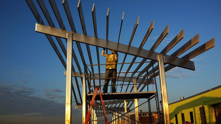 Show thumbnail preview A construction worker standing on scaffolding installs a modern, angular metal framework against a clear blue sky. The sun casts long shadows, highlighting the structures geometric design.