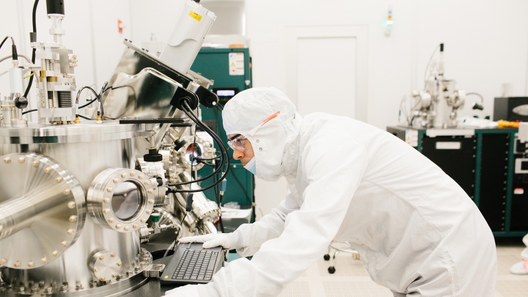 A scientist in a cleanroom suit operates a large, complex piece of laboratory equipment. They are bent over a computer keyboard, focusing intently. The room is bright and filled with high-tech machinery. A scientist in a cleanroom suit operates a large, complex piece of laboratory equipment. They are bent over a computer keyboard, focusing intently. The room is bright and filled with high-tech machinery.