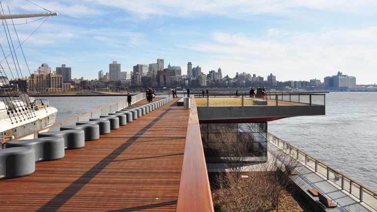 A waterfront boardwalk with modern seating extends toward a cityscape across the river. A large ship is partially visible on the left. People are scattered along the path, and the sky is bright with scattered clouds. A waterfront boardwalk with modern seating extends toward a cityscape across the river. A large ship is partially visible on the left. People are scattered along the path, and the sky is bright with scattered clouds.