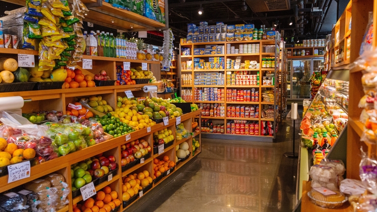 A well-organized grocery store aisle at Essex Market with shelves stocked with fresh fruits, vegetables, and various packaged goods. The produce section features a colorful array of oranges, apples, and bananas, while other shelves hold canned and boxed items. Photo by Kreg Holt/NYCEDC.