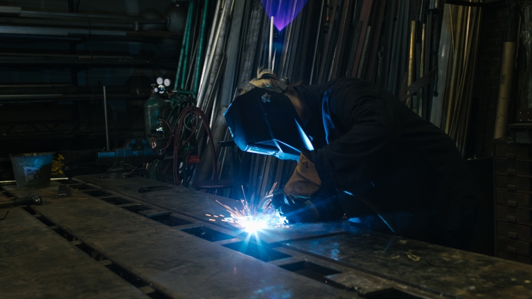 Person wearing a protective mask and gloves is welding metal in a dimly lit workshop, Makerspace. Sparks fly from the welding torch, illuminating nearby metal pipes and equipment. Photo Courtesy of Futureworks.