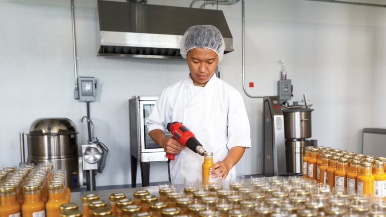 A MOMO Dressing employee in a white uniform and hairnet seals glass bottles of orange liquid with a heat gun in a food production facility. Numerous capped bottles are neatly arranged on a stainless steel table. Industrial kitchen equipment is visible in the background.