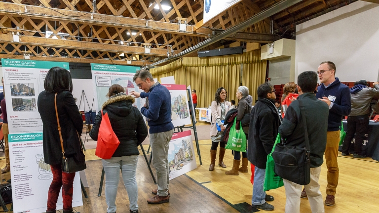 Show thumbnail preview A group of people in a community center are engaged in discussions, examining educational displays and posters on easels. Some hold reusable bags. The room has a wooden beamed ceiling and a stage with gold curtains.