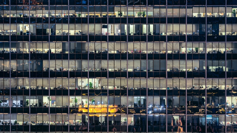 Night view of a high-rise office building with lit windows. Multiple floors are visible, showcasing office spaces with desks, computers, and scattered decor. The building reflects city lights and activity from within.