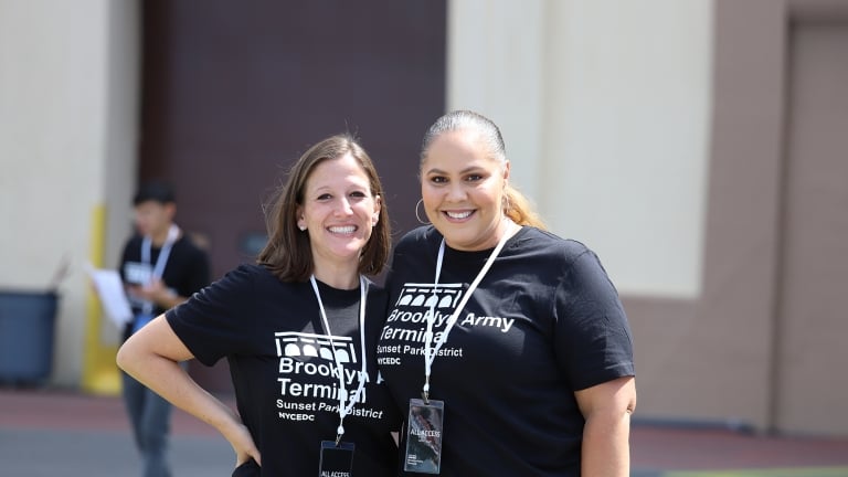 Two female NYCEDC employees at the BAT Block Party stand side by side outdoors, smiling at the camera. They are wearing matching black t-shirts and lanyards. A blurred background features a person walking and a building section.