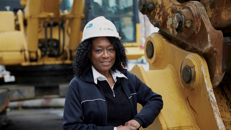 D'Ann Ricketts, Owner, BarDen Contracting Services, LLC, wearing a white hard hat and dark jacket stands confidently next to large yellow construction machinery. She is outdoors, with more equipment visible in the background. D'Ann Ricketts, Owner, BarDen Contracting Services, LLC, wearing a white hard hat and dark jacket stands confidently next to large yellow construction machinery. She is outdoors, with more equipment visible in the background.