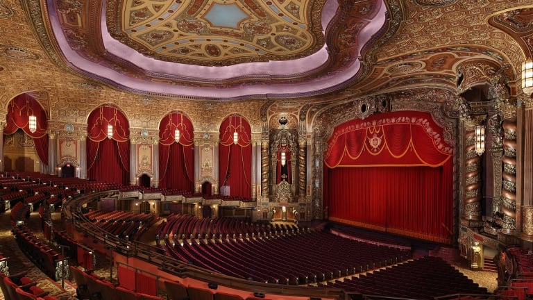 Ornate Kings Theatre interior with a grand ceiling featuring intricate designs and a large chandelier. Rows of red velvet seats face a stage with a huge red curtain. The walls are decorated with elaborate gold and red patterns. Photo by Whitney Cox/Courtesy of Kings Theatre.