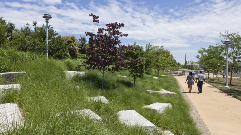 Photo of Hunts Point Landing courtesy of Matthew Nielsen Landscape Architects. A couple walks on a paved path in a park under a blue sky with scattered clouds. Lush green grass and scattered rocks are on the left, along with a variety of trees. Lamp posts line the path.