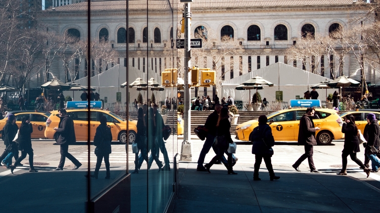 People walk past yellow taxis on a sunny day in a city. A large building with arches and a row of trees is in the background. A glass wall reflects the scene, adding symmetry and depth to the urban landscape.