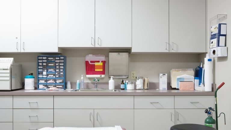 A medical examination room with white cabinets, countertop with various medical supplies, a sink, hand sanitizer, and a stool. An examination table with a paper cover is in the foreground. Various medical tools and protective gear are neatly arranged.